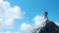 Businessman balancing on mountain peak against blue sky with clouds, symbolizing leadership challenges, career achievement and risk-taking in corporate world.