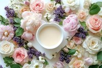 Cup of milk surrounded by delicate pink and white roses, peonies, and purple lilac flowers arranged on white background, creating romantic floral frame.