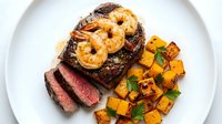 Grilled steak topped with caramelized pineapple rings, served with roasted sweet potato cubes and fresh herbs on white plate.
