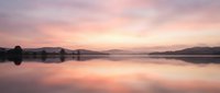 Serene lake at dawn with soft pink and purple sky reflecting on calm water surface, misty mountains in background, peaceful nature landscape.