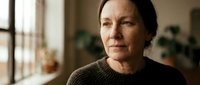 Middle-aged woman with dark hair wearing olive knit sweater indoors near window with natural light, contemplative expression, suitable for lifestyle and editorial use.