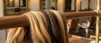 Long blonde hair with dark roots draped over wooden railing in warm-lit salon interior for beauty and haircare concepts.