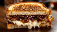 Grilled sandwich with melted cheese and caramelized onions on toasted bread, dripping with gooey cheese and golden-brown onions, served on wooden surface.