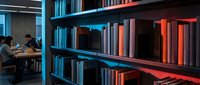 Modern library interior with organized bookshelves displaying colorful volumes in blue and red tones, two young students studying at wooden table in background, contemporary educational space.