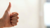 Hand showing thumbs up gesture against blurred background expressing approval, success and positive feedback in business or personal achievement.