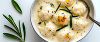 Creamy homemade dumplings in white sauce garnished with fresh rosemary sprigs and black pepper, served in ceramic bowl with vintage spoon.