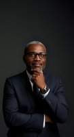 Middle-aged African American male professional wearing dark navy blazer and glasses, hand thoughtfully positioned near chin, contemplative expression against neutral background.