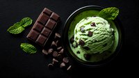 Mint chocolate chip ice cream scoop in dark bowl with fresh mint leaves and chocolate bar pieces scattered on black background, top view food photography.
