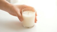 Hand holding a white soy wax candle in a glass jar on a light background, perfect for home decor, wellness, and aromatherapy concepts.