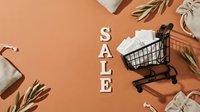 Miniature shopping cart filled with white cards beside wooden SALE letters on warm terracotta background with linen bags and olive branches.