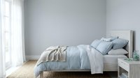 Serene bedroom interior with white bed frame, soft blue and beige bedding, natural light from window, minimalist decor, and wooden flooring for home design projects.