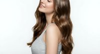 Young woman with long wavy brown hair in gray tank top smiling with eyes closed against white background. Hair care and beauty concept for salon advertising.