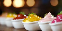 Colorful assorted dessert mousses served in white ceramic spoons, garnished with fresh herbs against blurred warm bokeh background, elegant catering setup.