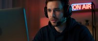 Male podcaster or streamer with dark hair and facial hair wearing headphones at desk with laptop in neon-lit studio setting, suitable for broadcasting, media, and entertainment content.