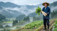 Middle-aged Asian male farmer in straw hat and blue shirt holding fresh harvested vegetables on hillside overlooking misty mountain valley with river and agricultural fields below.