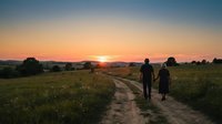 Two people walking along a rural dirt path through green meadow at golden hour sunset, with warm sky and distant trees creating serene countryside landscape.