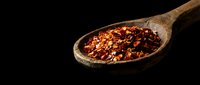Crushed red pepper flakes in wooden spoon on black background for culinary seasoning and spice cooking applications.