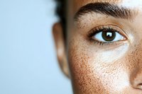 Close-up view of brown eye and natural freckles on light skin against blue background, showing authentic beauty and facial features in macro photography.