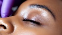 Young African American woman with closed eye showing professional makeup application with long eyelashes, shimmering eyeshadow and flawless skin texture.