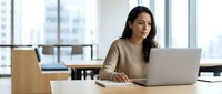 Young woman of South Asian descent working at laptop in modern office with city skyline view, suitable for business, technology, and corporate projects.
