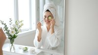 Young woman in white bathrobe applying cucumber eye patches for skincare routine in bright bathroom mirror reflection spa treatment at home.
