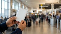 Traveler holding blank passport in airport terminal with crowds and departure boards visible in background, suitable for travel, immigration, and tourism concepts.