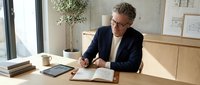 Mature man with gray hair and glasses wearing dark blazer and light shirt, writing in notebook at wooden desk with tablet and books in bright office space.