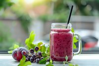 Fresh purple smoothie made with grapes and plums in mason jar with straw, garnished with mint leaves and fresh fruits on windowsill with natural light.