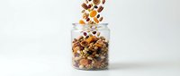 Mixed nuts and dried fruits pouring into glass jar on white background, isolated for food packaging, recipe blogs, nutrition content, and healthy snack marketing materials.