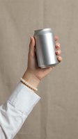 Female hand holding silver aluminum beverage can against neutral beige background. Minimalist product presentation with pearl bracelet accent for branding mockup.