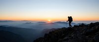 Young adult male hiker with backpack standing on rocky mountain summit at sunrise, overlooking misty valleys and distant peaks. Adventure and outdoor exploration concept.