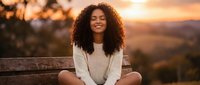 Young African American woman with curly hair sitting peacefully on wooden bench during golden hour sunset, eyes closed in meditation and mindfulness moment.