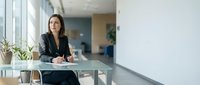 Professional businesswoman in black suit sitting at modern office desk with laptop and documents in bright contemporary workspace with large windows.