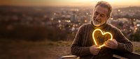 Senior Caucasian man holding glowing heart-shaped light at sunset overlooking city skyline. Romantic gesture, love concept for Valentine's Day greeting cards.
