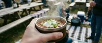 Hand holding wooden bowl of hot soup with steam rising at outdoor food festival or market event with blurred crowd background for culinary dining concepts.