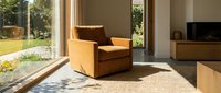 Warm-toned upholstered armchair positioned in bright living room with large windows, natural light, wooden furniture, and garden view for interior design and home decor.