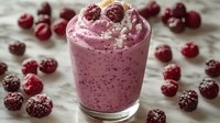 Creamy raspberry smoothie topped with powdered sugar in glass, surrounded by fresh frozen berries on white marble surface. Healthy breakfast drink.