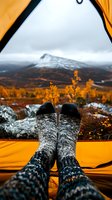 Cozy wool socks inside camping tent with view of snow-capped mountain and autumn landscape through tent opening, creating warm outdoor adventure atmosphere.