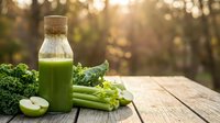 Fresh green juice in glass bottle with cork surrounded by kale, celery, apple slices on wooden table with warm sunlight bokeh background for healthy lifestyle.
