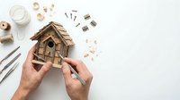Caucasian male hands assembling a rustic wooden birdhouse on white background, with tools, screws, and twine nearby.