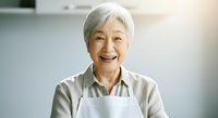 Senior woman with white hair, East Asian ethnicity, wearing white apron over beige shirt, smiling warmly in bright kitchen setting. Suitable for culinary, cooking, hospitality, and lifestyle content.