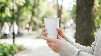 Person holding blank white disposable cup in park setting with green trees and natural daylight, suitable for beverage branding, coffee shop marketing, and eco-friendly packaging mockups.