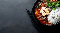 Spicy Korean tofu stew with steamed rice, sesame seeds, and fresh herbs in a black bowl with chopsticks on dark stone background.