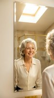 Happy senior Caucasian woman with gray hair smiling at her reflection in bathroom mirror during morning routine in bright natural light from skylight.