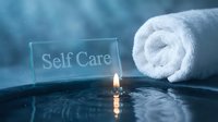 Glowing candle beside a rolled white towel and glass sign reading Self Care on a reflective surface in soft blue tones.