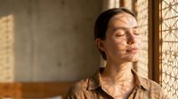 Woman with dark hair and olive complexion wearing tan shirt gazes thoughtfully through sunlit window with geometric pattern, conveying contemplation and peaceful solitude in minimalist interior space.