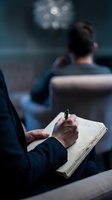 Businessman writing notes in notebook during corporate meeting or conference with blurred colleagues in background for business planning and strategy.