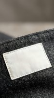 Blank white fabric label sewn on dark gray wool textile with stitched edges for branding and customization purposes in fashion and apparel manufacturing.