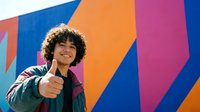 Young man with curly hair and mixed ethnicity wearing casual jacket pointing at camera against vibrant geometric background with blue, orange, and pink stripes for advertising and marketing campaigns.