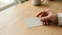 Young adult male with light skin tone holding blank white business card on wooden desk with coffee mug, promoting corporate branding and professional networking opportunities.